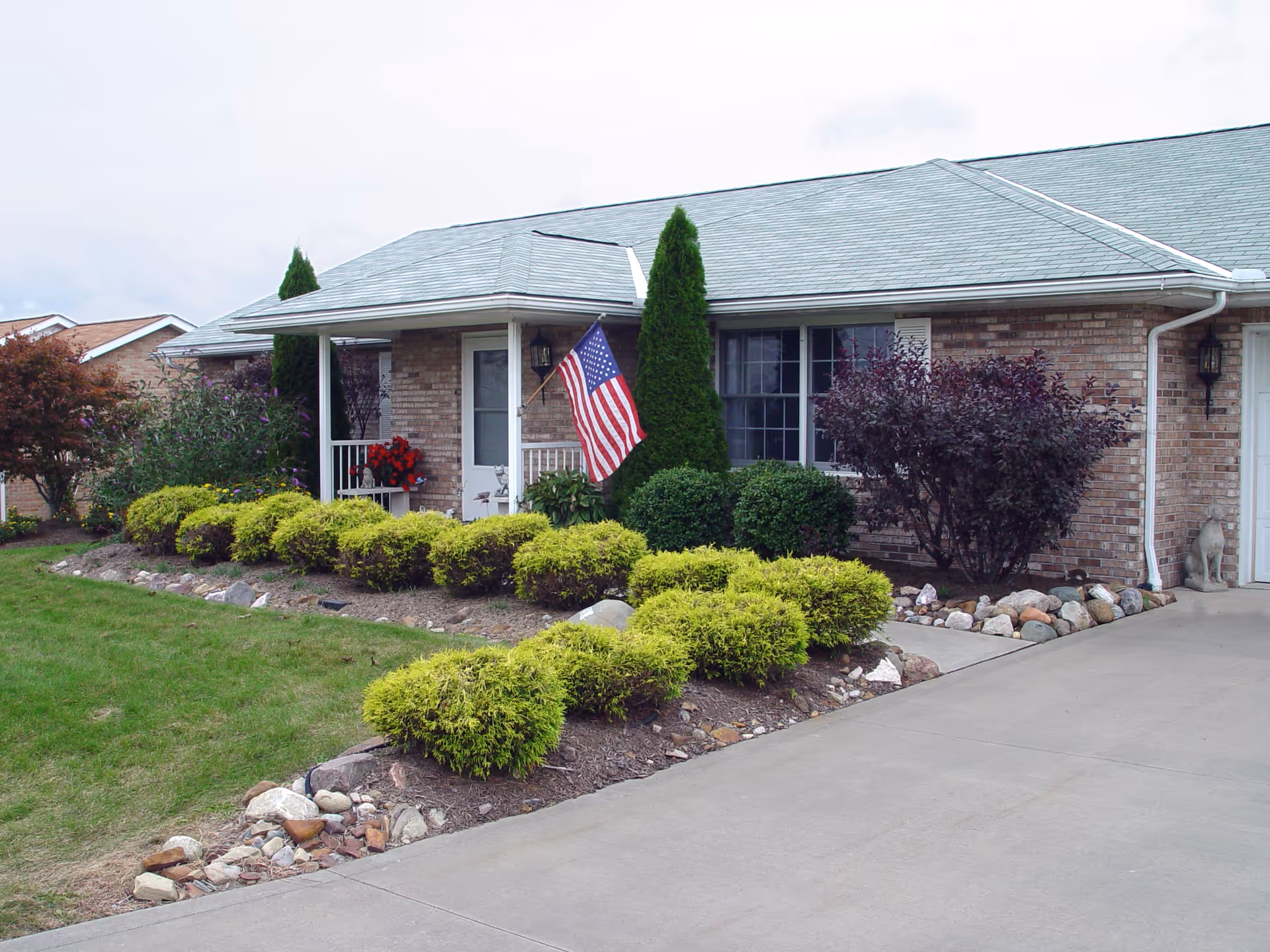 Exterior view of a single-story brick building with a gray roof, a small porch with white railings, an American flag mounted near the entrance, and well-maintained landscaping including green bushes, shrubs, and a tree. A concrete driveway is visible in the foreground.