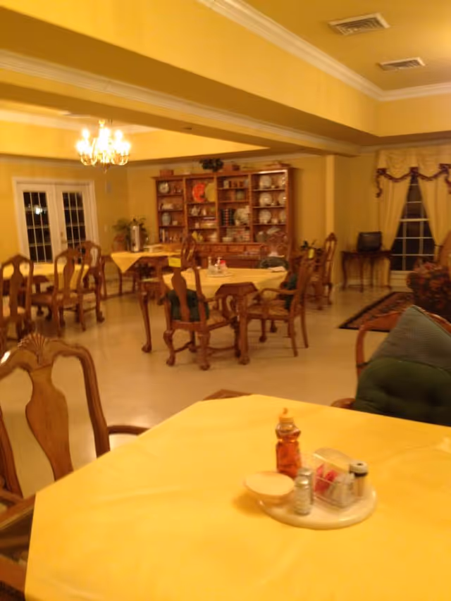 A dining room with several wooden tables and chairs, each table covered with a yellow tablecloth. The room has yellow walls and a chandelier hanging from the ceiling. In the background, there is a wooden cabinet filled with plates and decorative items. A window with yellow curtains is visible on the right side.