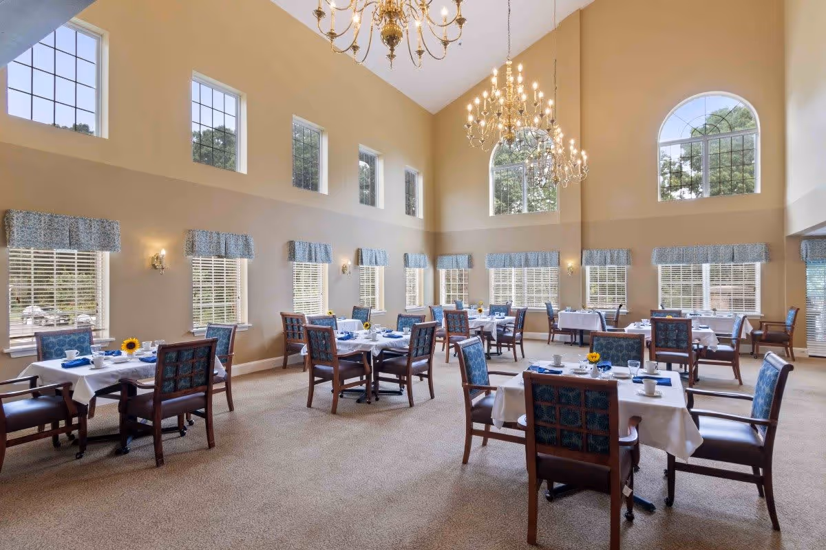 A spacious dining room with high ceilings and large windows letting in natural light. The room is furnished with multiple tables covered with white tablecloths, each set with blue napkins, cups, and small vases holding sunflowers. Elegant chandeliers hang from the ceiling, and the walls are painted a warm beige color.