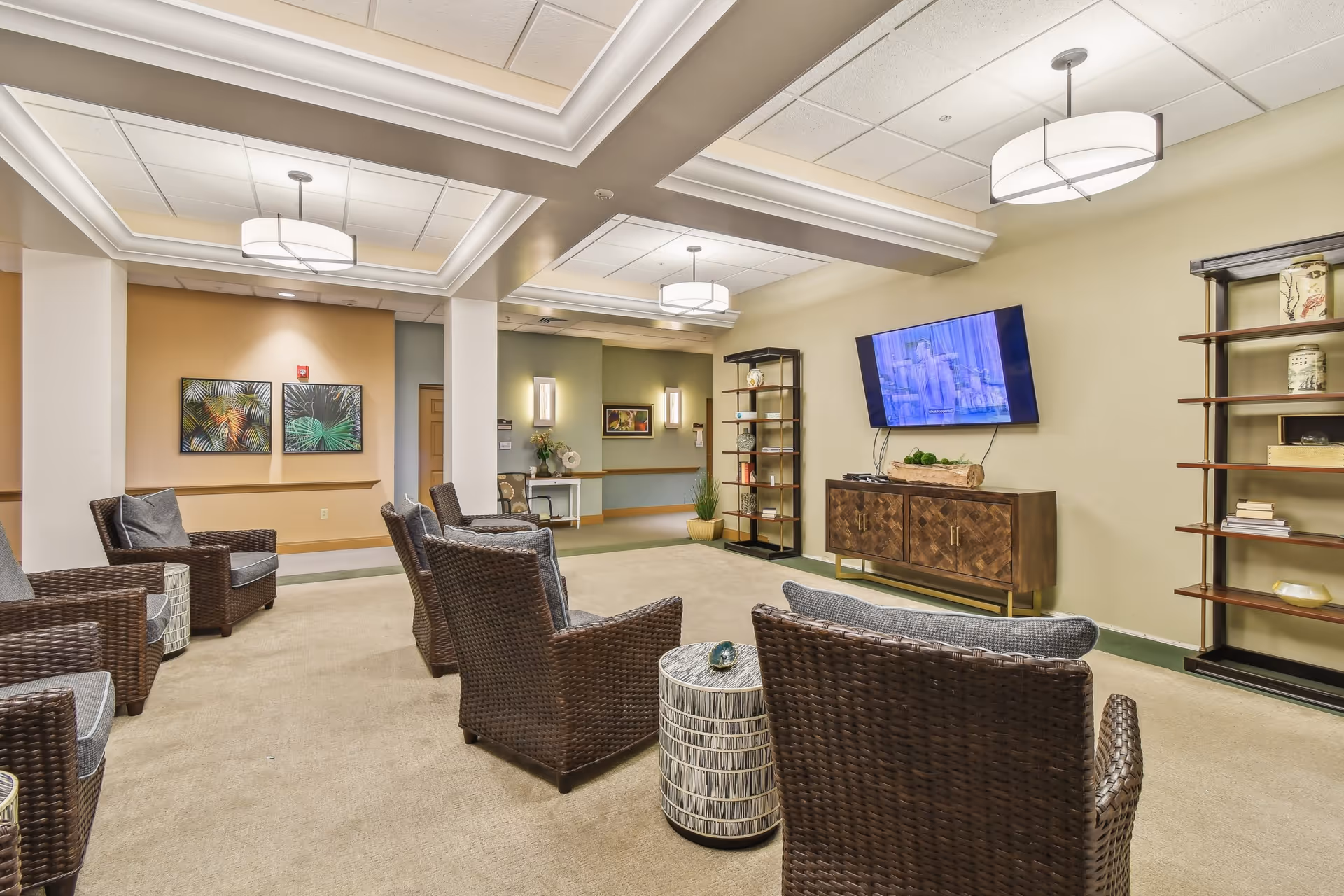 A cozy lounge area with multiple wicker armchairs arranged facing a wall-mounted flat screen TV. The room features a beige carpet, soft yellow and green walls, decorative shelving units with vases and books, and ceiling lights with modern fixtures. Two framed botanical prints hang on the far wall.