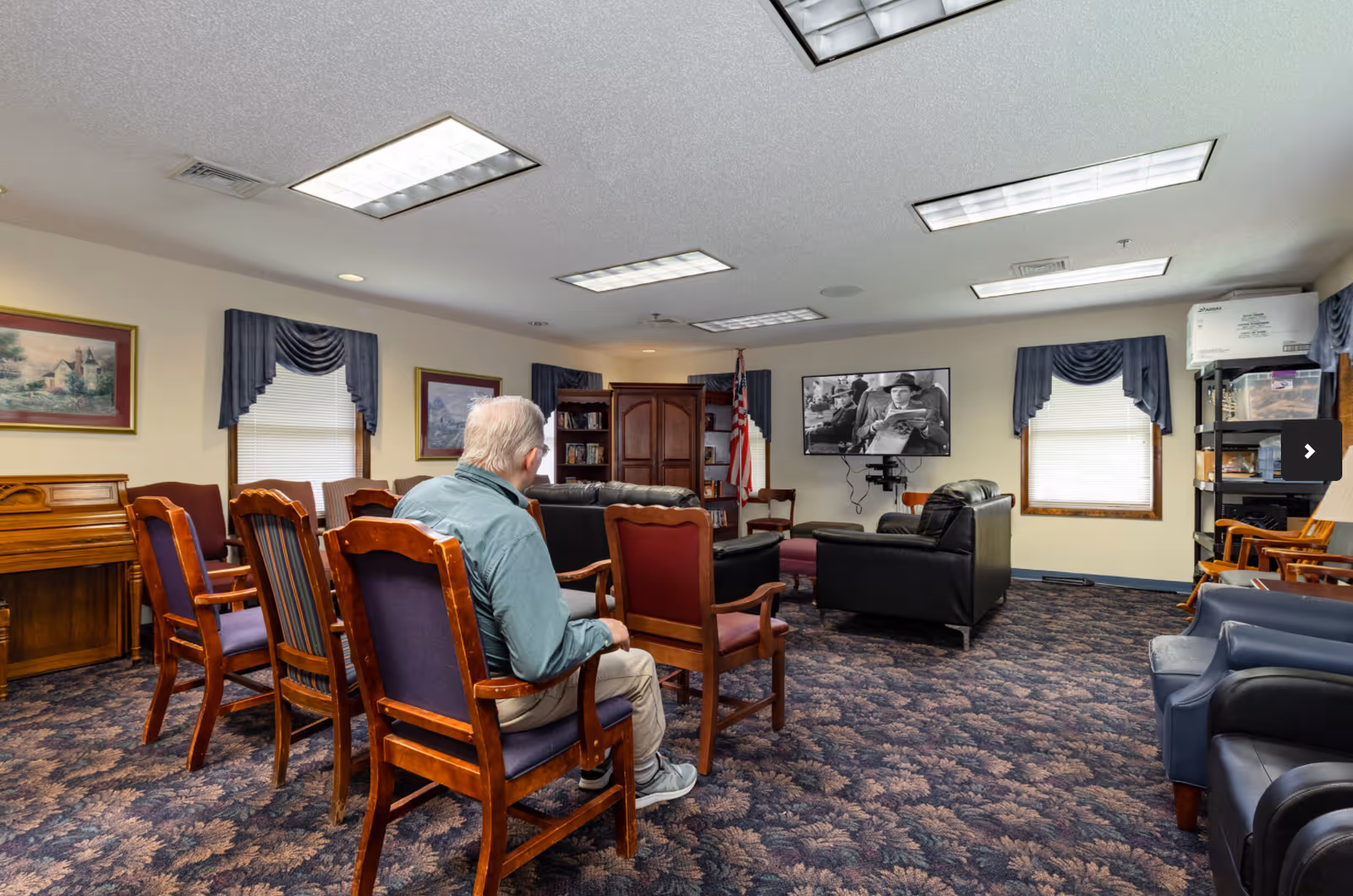 A senior man sitting on a wooden chair in a common living room area with multiple chairs, a piano, and a television mounted on the wall showing a black and white movie. The room has patterned carpet, framed paintings on the walls, windows with blue valances, and fluorescent ceiling lights.