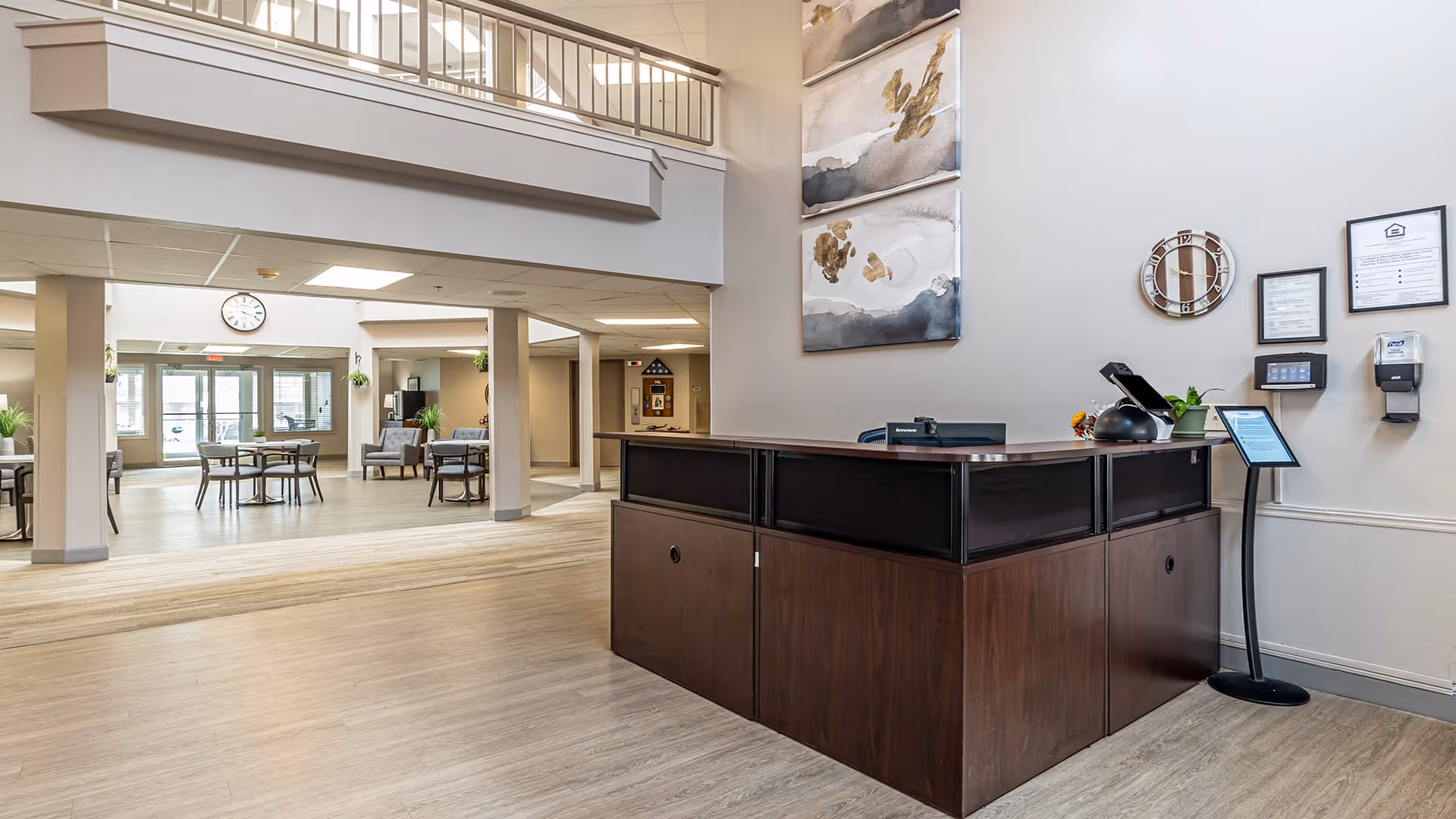 Reception area of Holiday Quail Run Estates featuring a dark wood front desk with a computer, phone, and decorative items. Behind the desk are two abstract paintings and a round mirror on the wall. The space has light-colored walls and flooring, with an open view into a seating area with tables and chairs, and large windows letting in natural light.