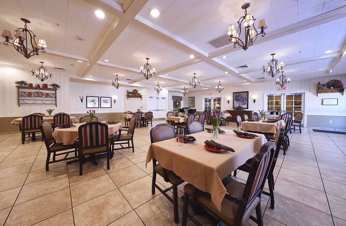 A spacious dining room with multiple tables covered in beige tablecloths, each set with plates, glasses, and napkins. The room features tiled floors, decorative chandeliers hanging from a coffered ceiling, and walls adorned with framed artwork and shelves with decorative items. Chairs with striped upholstery surround the tables.