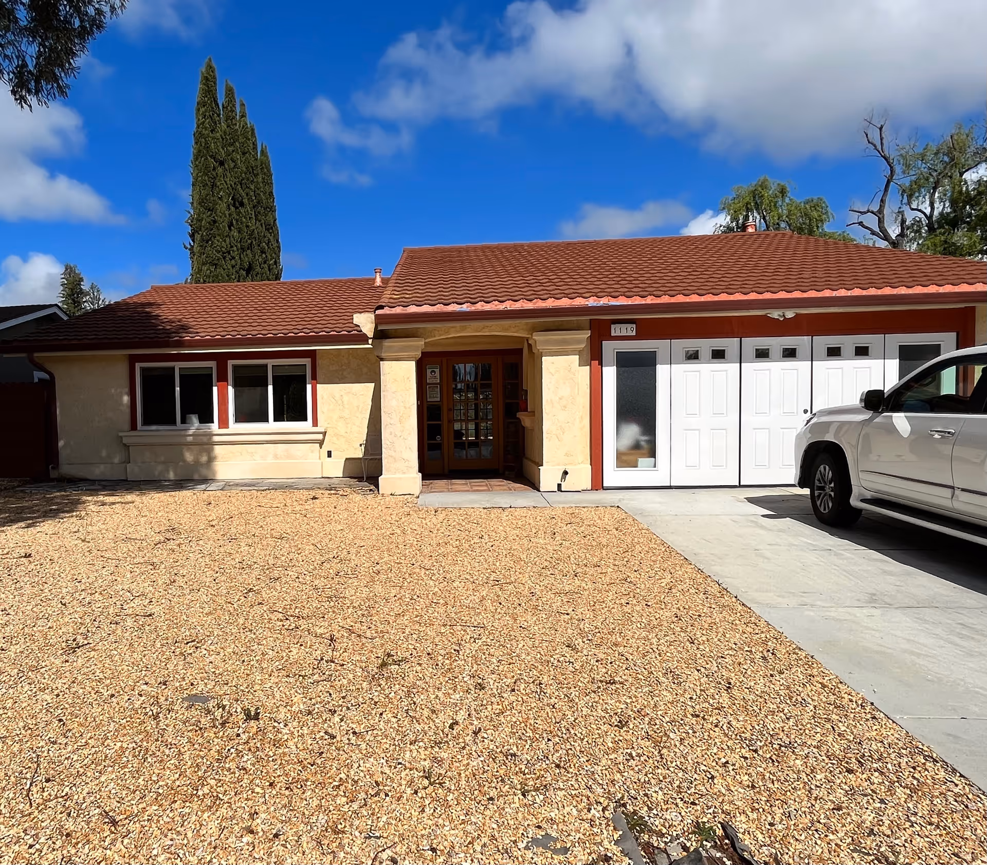 Front exterior view of a single-story house with a red-tiled roof, beige stucco walls, and a driveway with a white SUV parked. The front entrance has a small covered porch with two columns, and there are three windows on the left side of the house. The yard is covered with gravel and there are tall trees in the background under a blue sky with some clouds.