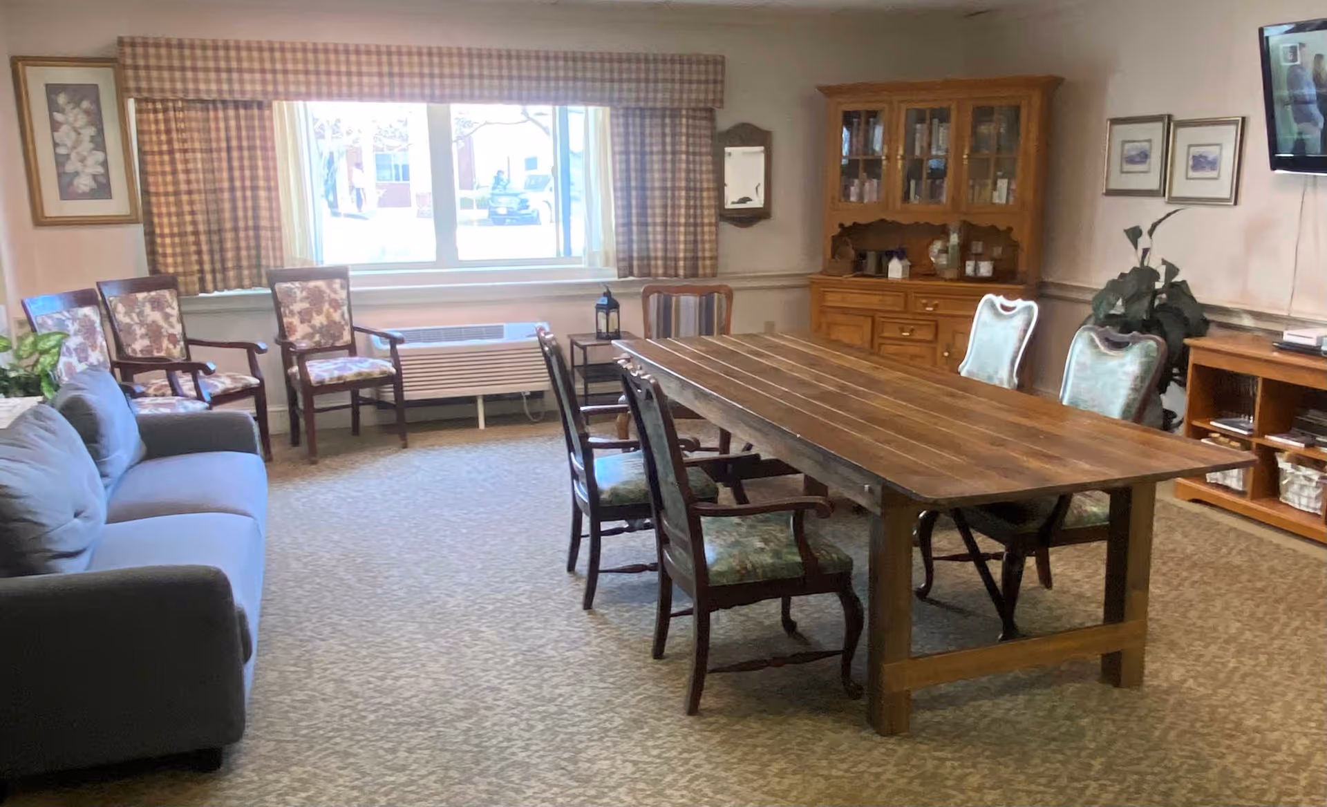 A cozy common room with a large wooden table surrounded by six chairs with floral upholstery. There is a gray sofa on the left side and three additional floral armchairs near a window with plaid curtains. A wooden cabinet with glass doors and a TV mounted on the wall are visible on the right side. The room is carpeted and decorated with framed pictures and a potted plant.