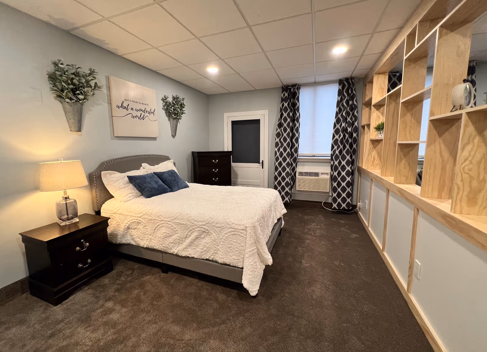 Cozy bedroom with a queen bed dressed in white bedding, a nightstand and lamp, dresser, window with patterned curtains, and built-in wooden shelving.