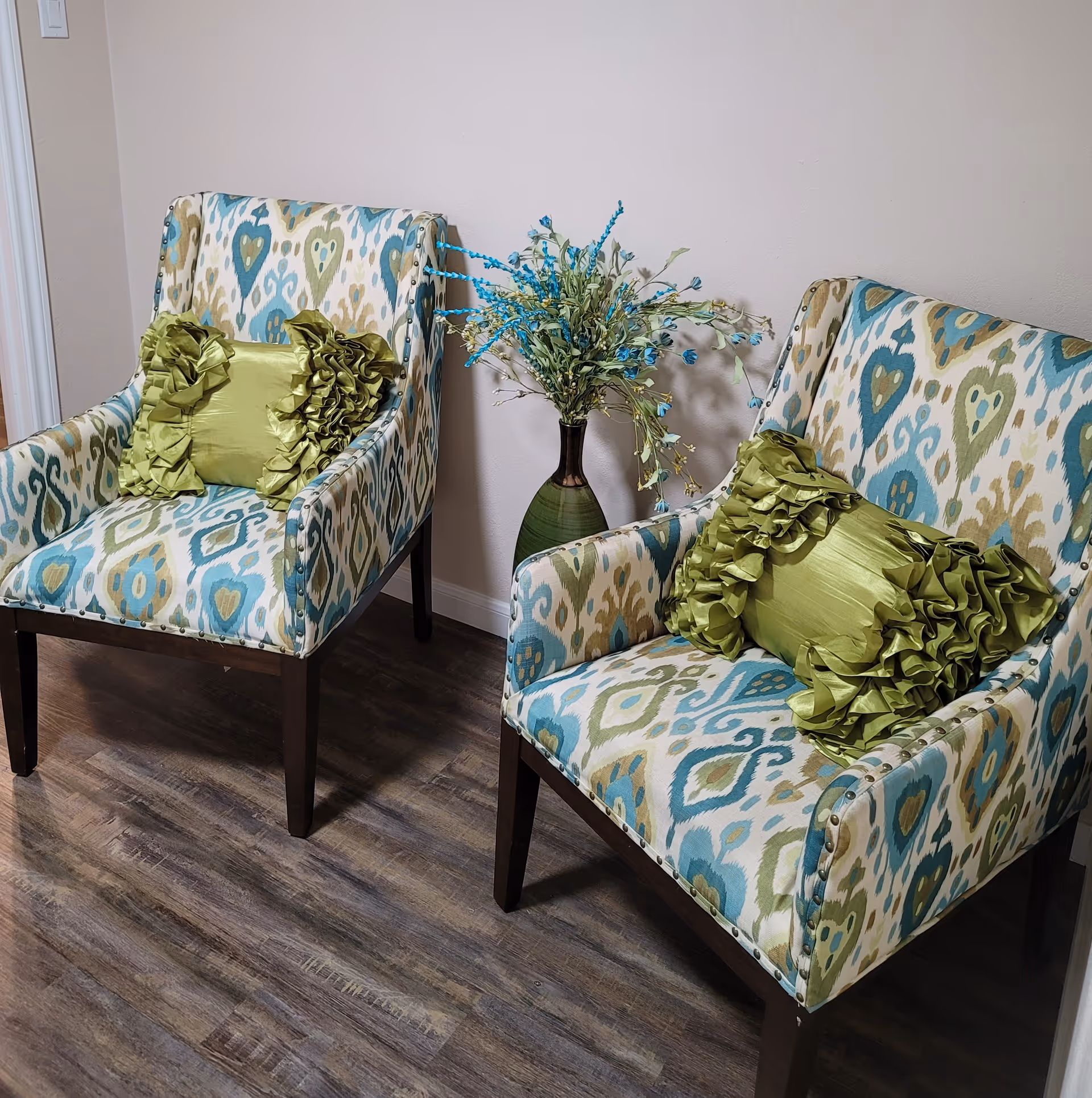 Two patterned upholstered armchairs with green ruffled pillows flanking a vase of blue-green flowers on a wood floor against a pale wall.