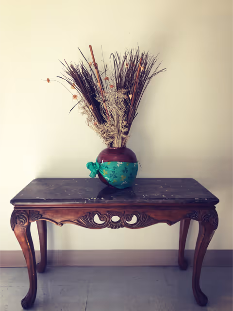 A decorative wooden table with carved details and curved legs, topped with a dark marble surface. On the table is a round vase with a green and brown design, holding dried ornamental grasses and twigs. The background is a plain light-colored wall and a tiled floor.