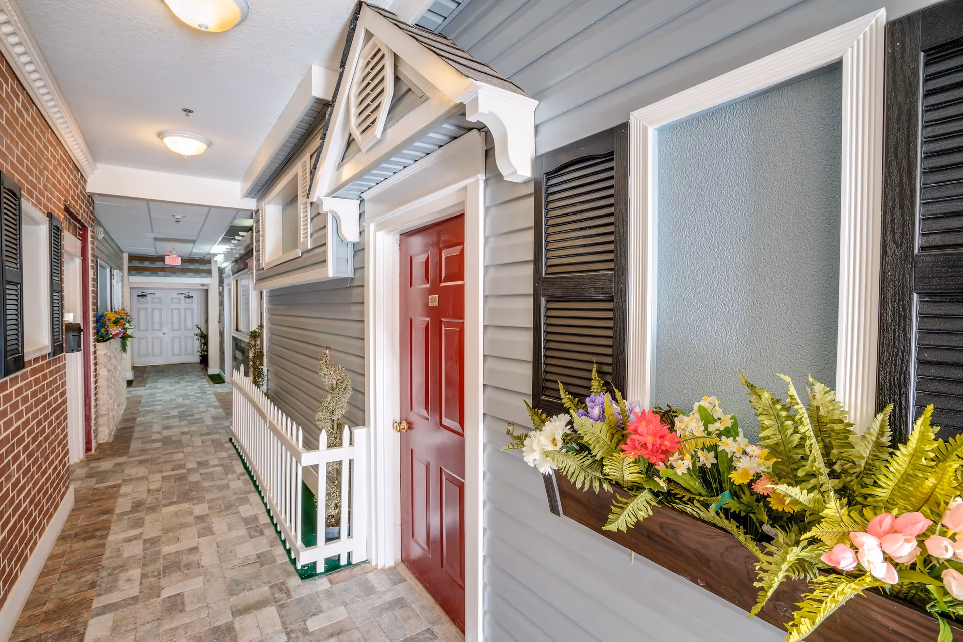 Indoor hallway designed to look like an outdoor street with house facades, including a red door labeled 308, window shutters, flower boxes with colorful flowers, a white picket fence, and brick and siding walls under ceiling lights.