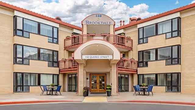 Front exterior view of Brookdale Lowry building with beige brick walls, large windows, and a central entrance under an archway labeled '150 Quebec Street'. There are outdoor tables and chairs on either side of the entrance, with potted plants near the doorway. The sky is partly cloudy.