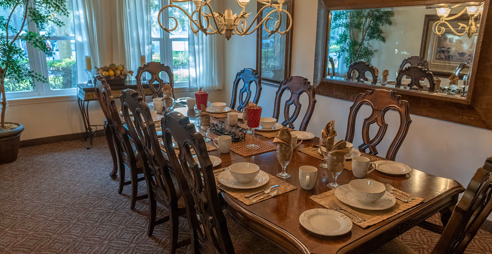 A formal dining room with a long wooden table set for eight people. The table is arranged with white plates, bowls, cups, silverware, and red decorative goblets. The room has large windows with sheer white curtains, a large mirror on one wall, a chandelier overhead, and potted plants near the windows.
