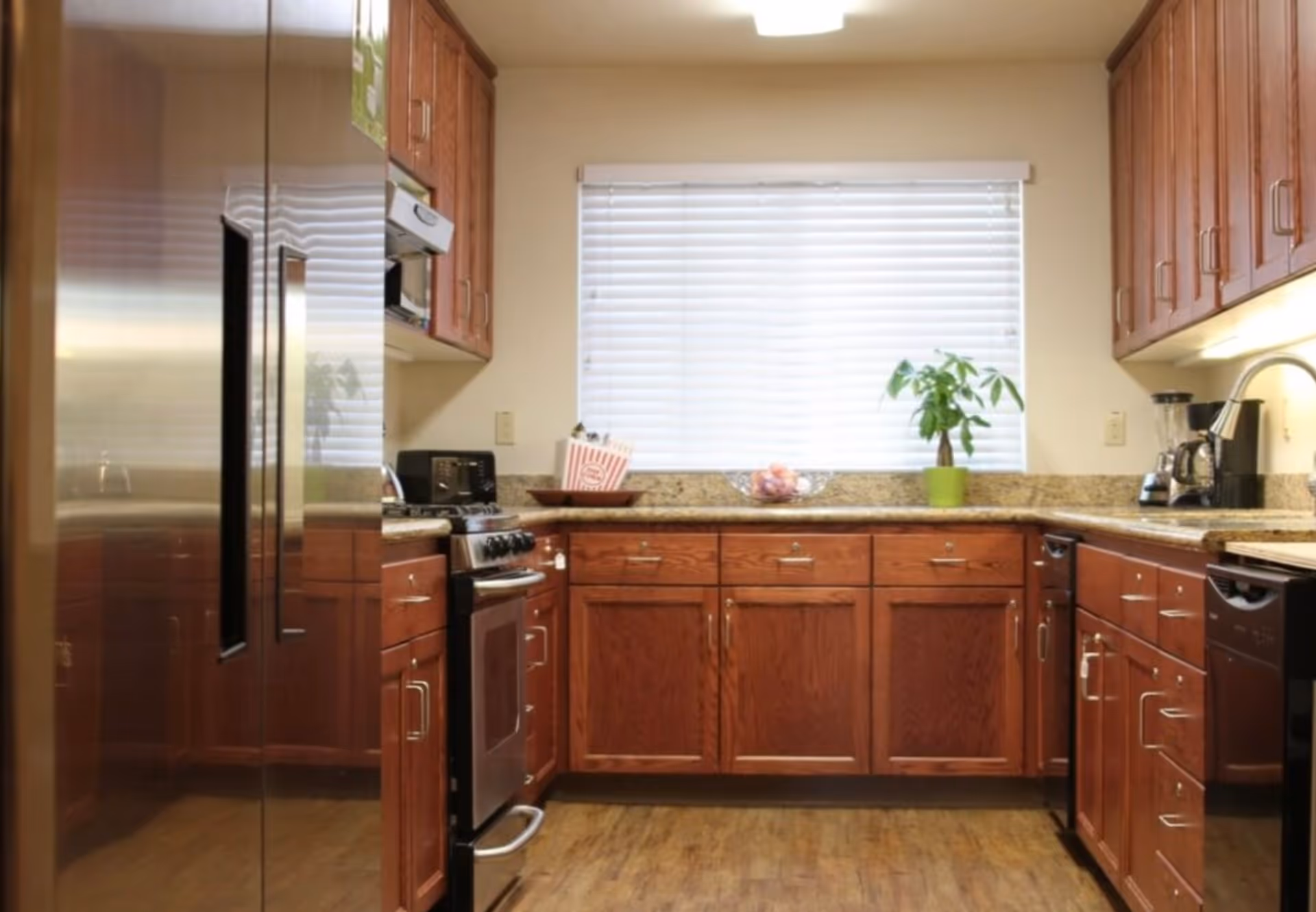 A kitchen with wooden cabinets, granite countertops, a stainless steel refrigerator, a stove, a microwave, a toaster, a coffee maker, a blender, and a window with white blinds. There is a small green potted plant and a bowl of fruit on the countertop.