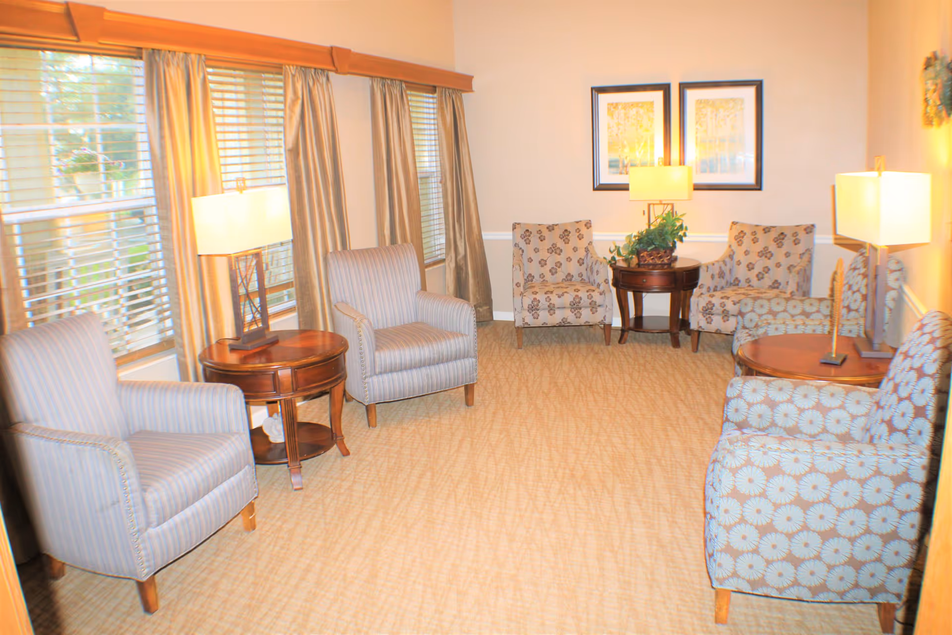 A cozy sitting room with six upholstered armchairs arranged around three wooden side tables with lamps. The room has beige carpeting, cream-colored walls, large windows with blinds and gold curtains, and two framed pictures on the wall.