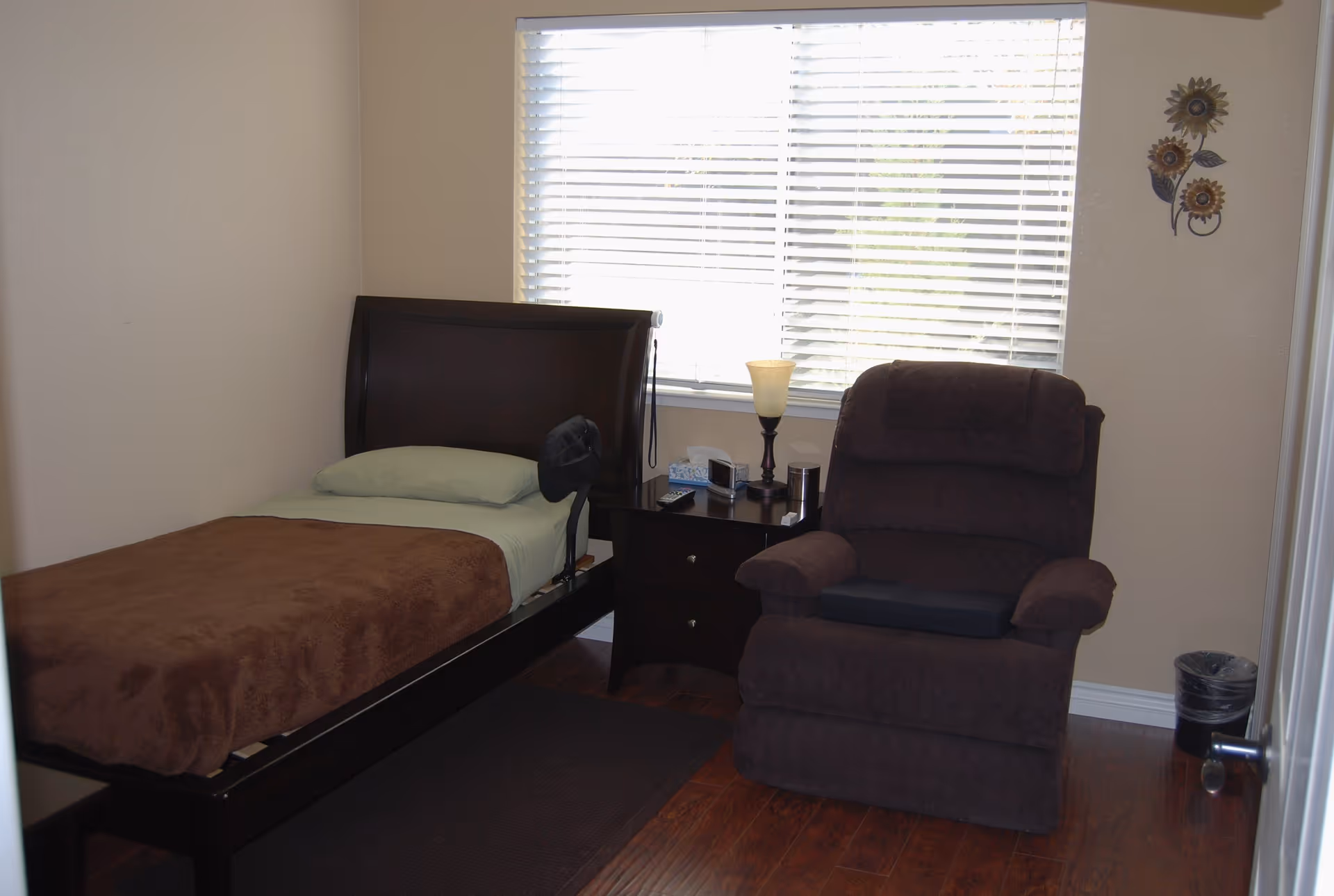A small bedroom with a single bed covered with a brown blanket and a light green pillow. Next to the bed is a dark wooden nightstand with a lamp, tissue box, and other small items. A large brown recliner chair is positioned beside the nightstand. The room has wooden flooring, beige walls, a window with white blinds, and a decorative wall hanging with three metal flowers.