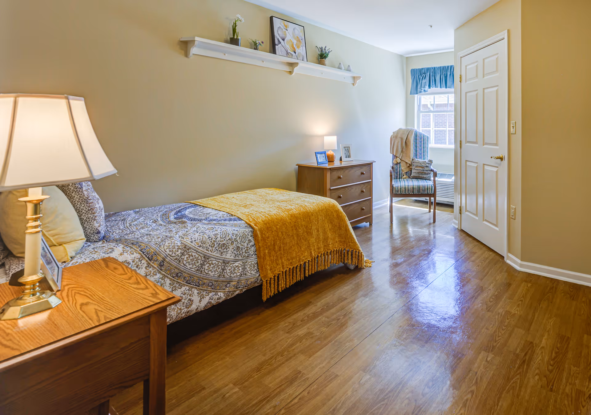 A neatly arranged bedroom with a single bed covered in patterned bedding and a mustard yellow throw blanket. There is a wooden nightstand with a lamp and a small picture frame next to the bed. Across from the bed is a wooden dresser with a lamp, picture frames, and a small plant. A chair with a blanket draped over it is positioned near a window with blue curtains. The room has wooden flooring and light-colored walls with a decorative shelf holding framed pictures and small plants.