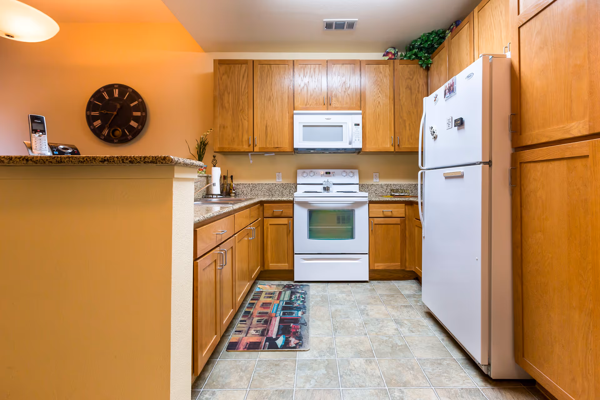 A kitchen with wooden cabinets, a white refrigerator, a white stove with an oven, and a white microwave above the stove. There is a granite countertop with a sink and a decorative mat on the tiled floor. A wall clock and a cordless phone are visible on the left side.