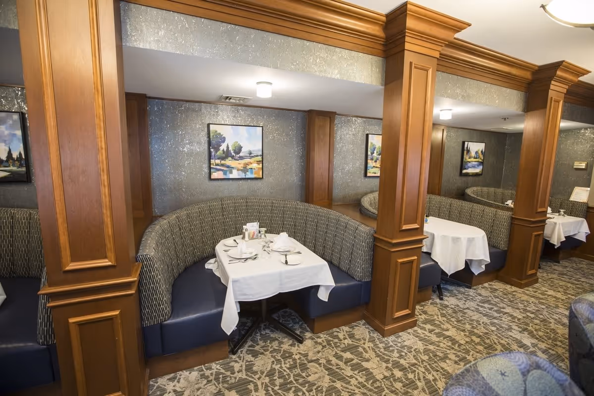 Interior view of a dining area with curved booth seating and tables set with white tablecloths, plates, and napkins. The walls have a textured finish and are decorated with framed landscape paintings. Wooden columns separate the booths, and the floor is carpeted with a patterned design.