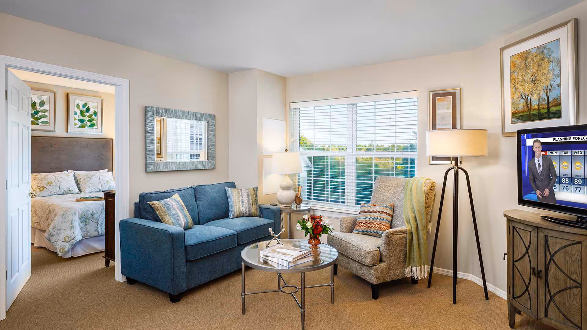 A cozy living room with a blue sofa, a beige armchair with a throw blanket, a round glass coffee table with books and a flower vase, a floor lamp, and a TV showing a weather forecast. The room has a large window with blinds and light-colored walls. Through an open doorway, a bedroom with a bed, floral bedding, and framed leaf artwork is visible.