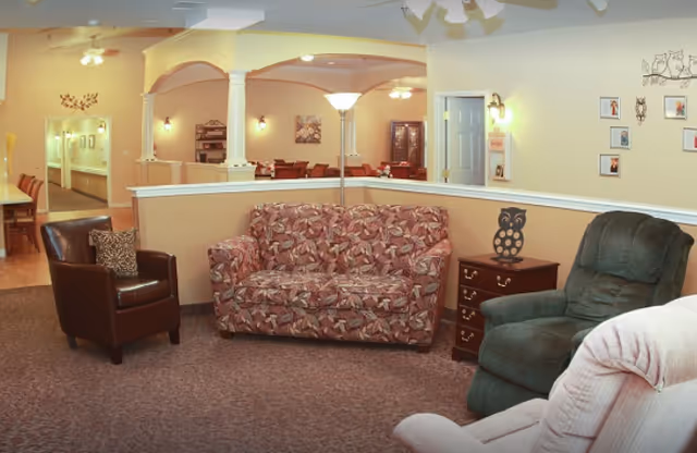 Cozy senior living common area with a patterned loveseat, several armchairs, a side table, and arched openings to a dining space.