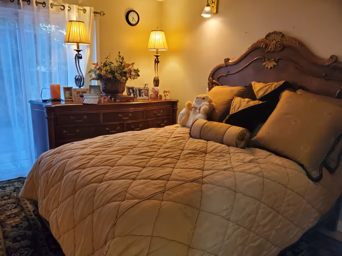 A cozy bedroom with a large wooden bed featuring an ornate headboard, covered with a quilted beige bedspread and multiple pillows. A stuffed animal is placed on the bed. Next to the bed is a wooden dresser with two lit table lamps, framed photos, and a flower arrangement. A clock is mounted on the wall above the dresser, and sheer curtains cover a window or glass door on the left side.