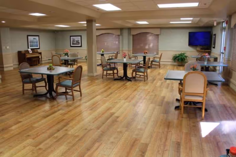 Spacious communal dining/activity room with several tables and chairs, a piano, and a wall-mounted TV.