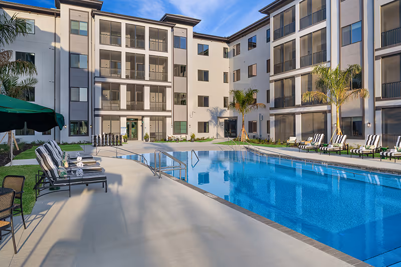 Sunlit courtyard featuring a rectangular swimming pool, striped lounge chairs, and a multi-story residential building.