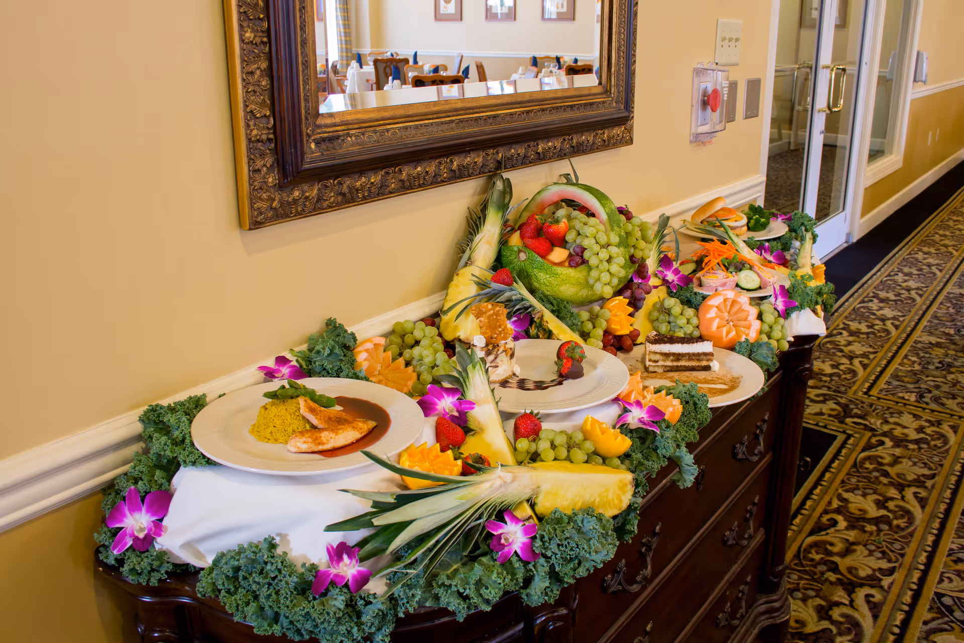 A decorative buffet table with plates of food including grilled chicken with rice, desserts, and an assortment of fresh fruits such as grapes, strawberries, pineapple, and carved melons, garnished with purple flowers and leafy greens. A large ornate mirror hangs on the wall above the table, and a dining area with tables and chairs is visible in the reflection.