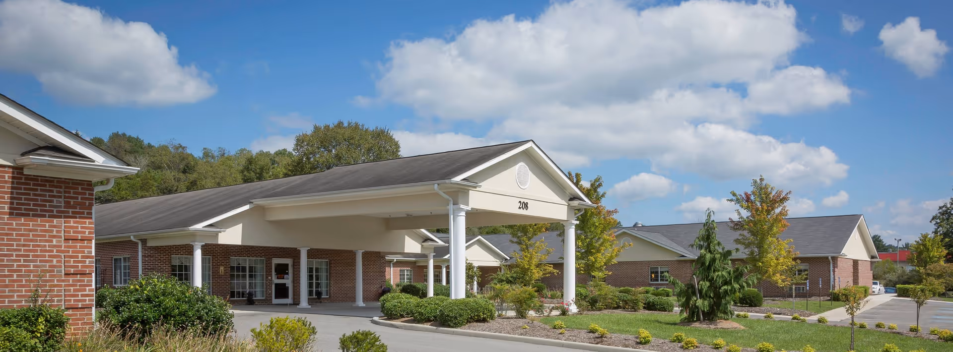Exterior view of a single-story brick building with a covered entrance supported by white columns, surrounded by landscaped greenery and trees under a partly cloudy blue sky.