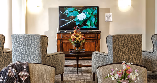 A cozy living room area with patterned armchairs arranged around a wooden coffee table with a floral centerpiece. A wooden cabinet holds a flat-screen TV displaying an image of green leaves and white flowers. The room has neutral-colored walls and soft lighting.