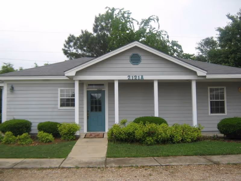Front exterior of a single-story gray house with a teal entrance door, porch columns, shrubs, and address numbers above the door.