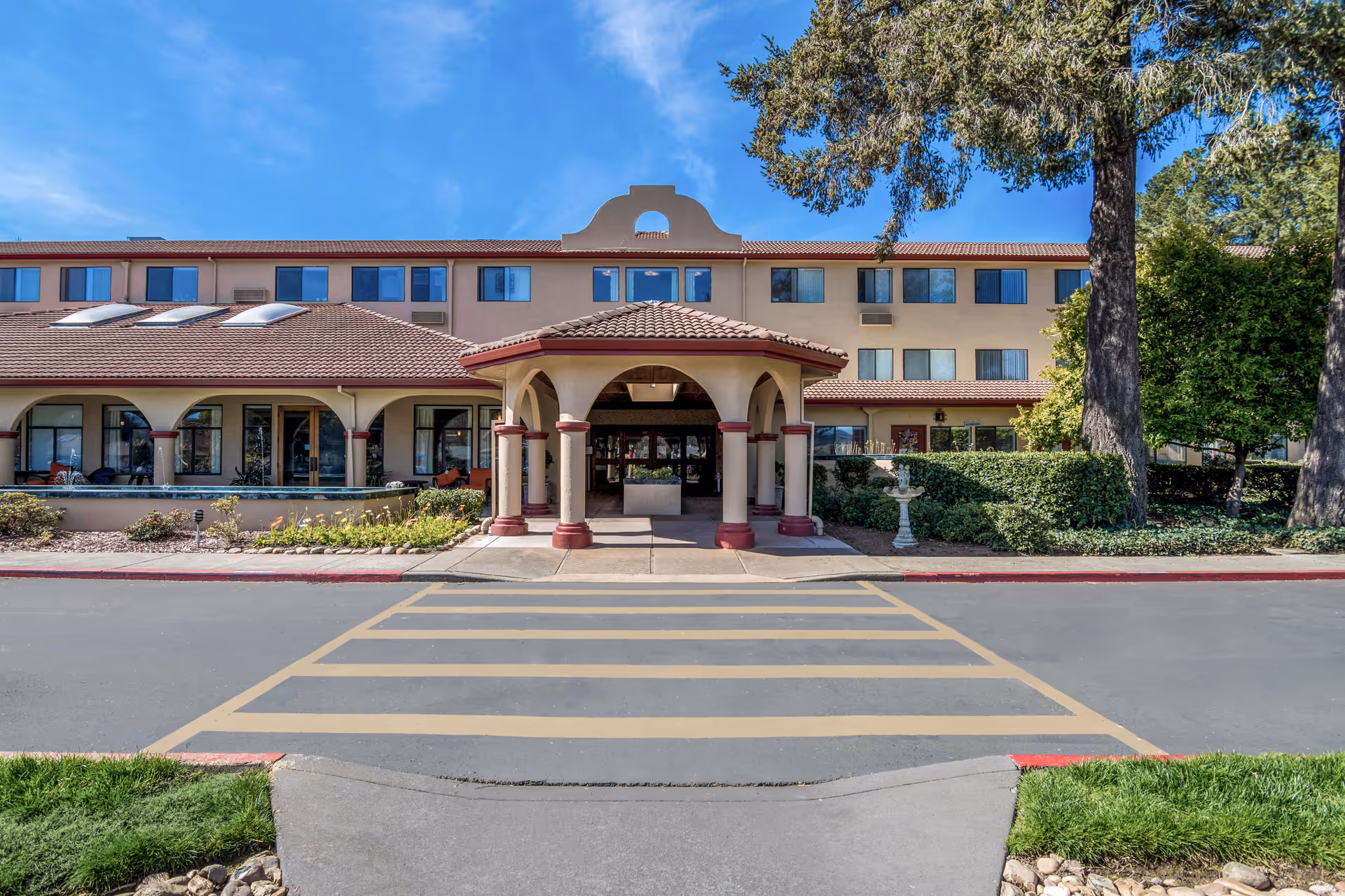 Front exterior view of Holiday Redwood facility showing a three-story building with a covered entrance, arched windows, and a tiled roof under a blue sky with some trees and landscaping around.