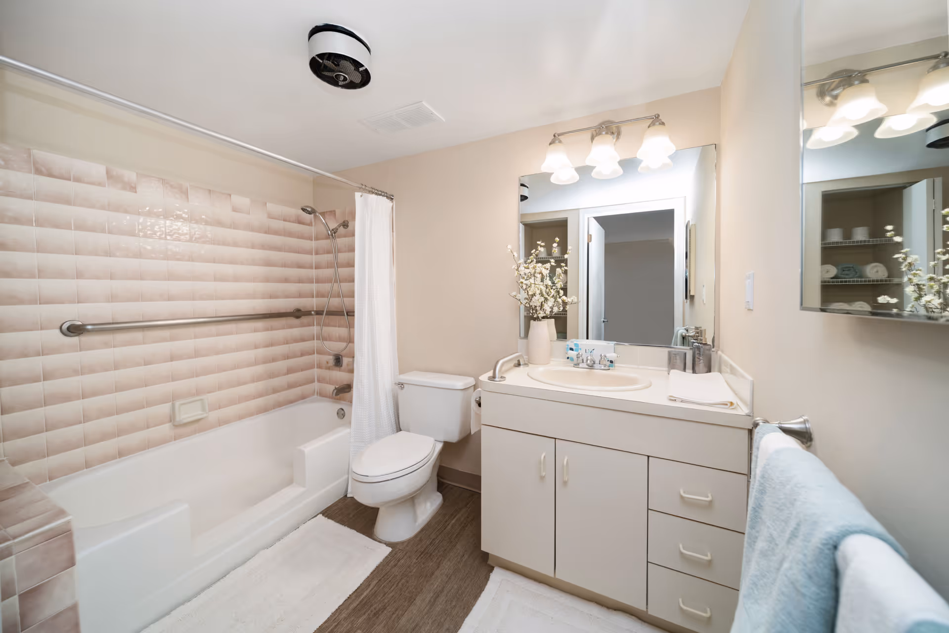 Well-lit bathroom with a bathtub/shower, grab bar, toilet, and a vanity with mirror and decorative flowers.