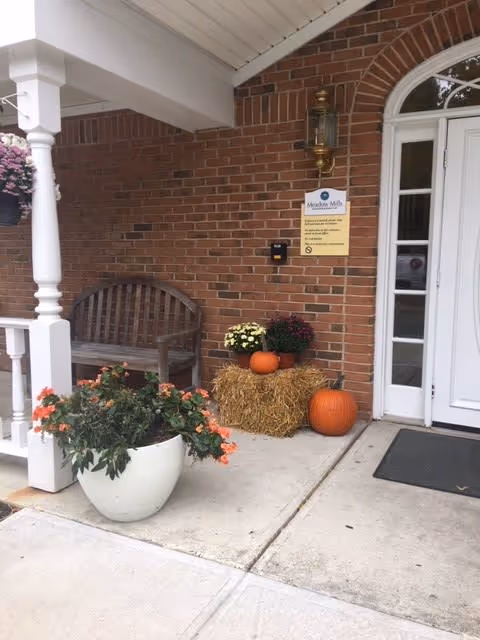 Entrance area of Meadow Mills At Whitney Center featuring a brick wall, a white door with glass panels, a wooden bench, a white planter with orange flowers, a bale of hay decorated with two pumpkins and two pots of flowers, and a wall-mounted lantern light.