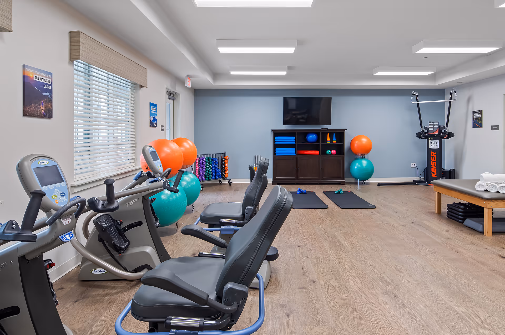 A bright fitness room with recumbent exercise bikes, stability balls, mats, small weights and a TV cabinet.
