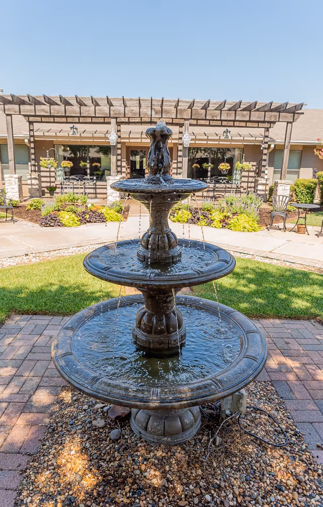 A three-tiered water fountain in the center of a landscaped outdoor area with a brick walkway and green grass. In the background, there is a building with large windows, a wooden pergola, hanging flower baskets, and outdoor seating.