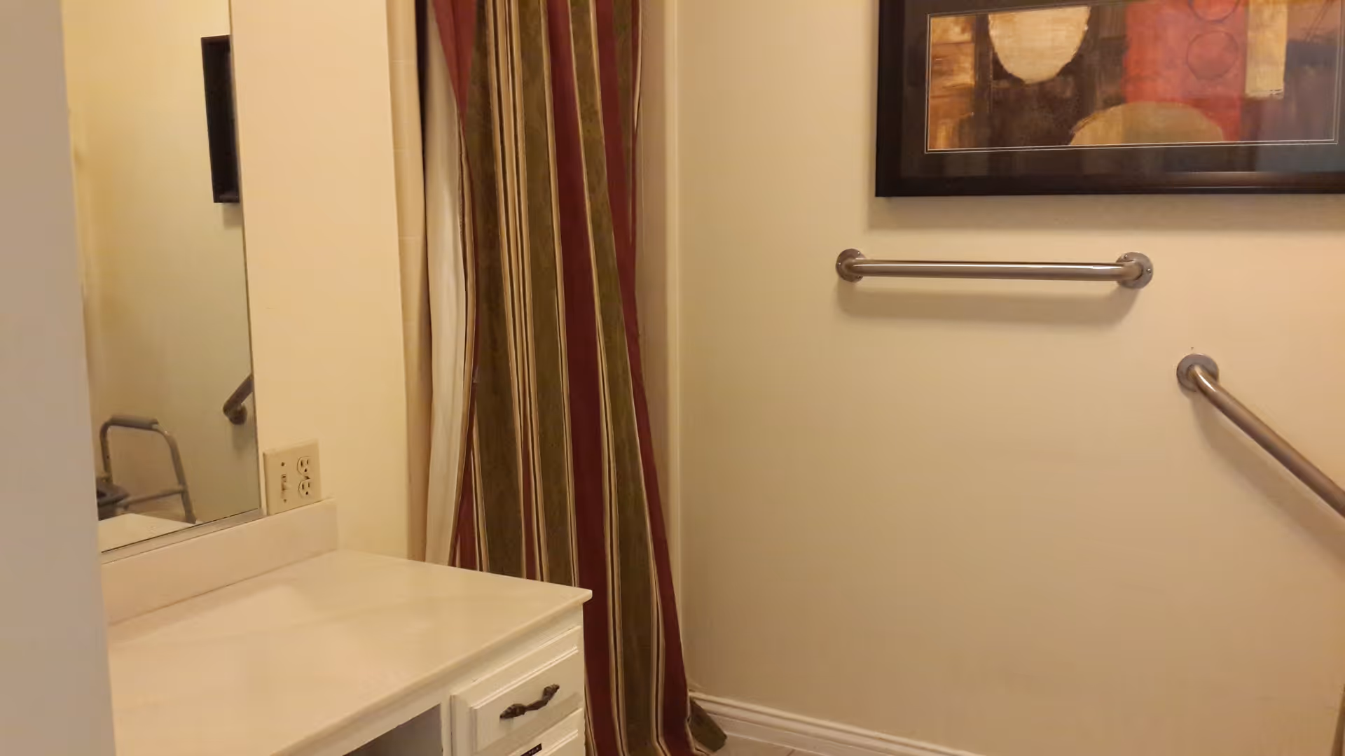 Interior view of a bathroom with a white countertop and cabinet, a large mirror, grab bars on the wall, a striped shower curtain, and a framed abstract painting above the grab bars.