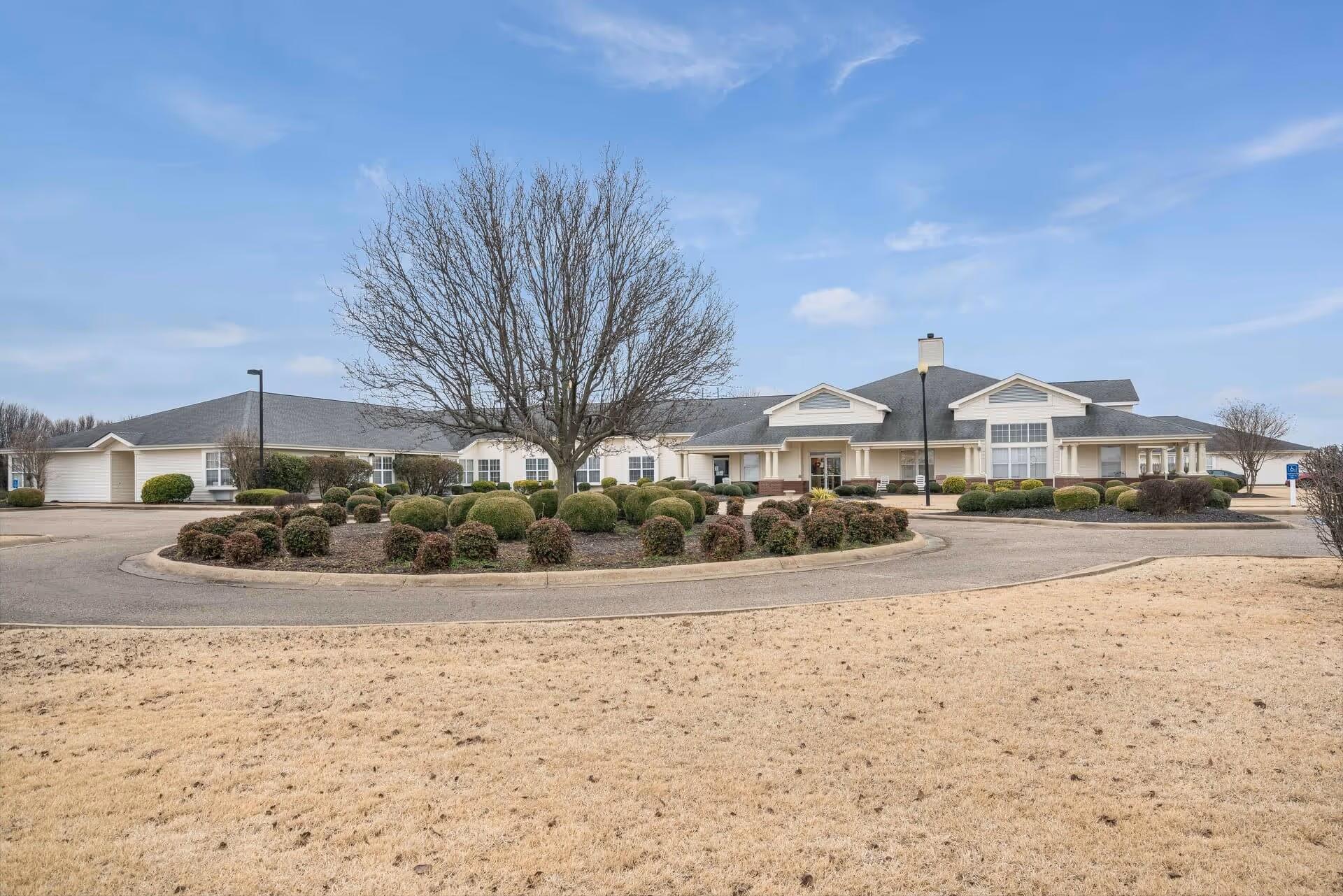 Exterior view of a single-story senior living facility building with a circular driveway and landscaped bushes and trees in front. The sky is clear with a few clouds.