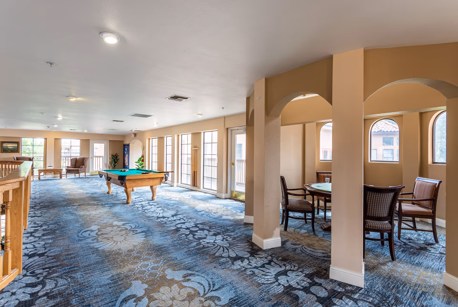 A spacious senior living common area with a pool table in the center, a seating area with armchairs and a coffee table near large windows, and a separate nook with a round table and four chairs under arched openings. The room has patterned blue carpet and beige walls with multiple windows letting in natural light.