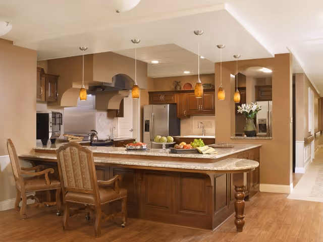 A spacious kitchen area with a large granite island countertop featuring wooden cabinetry and three upholstered chairs. The kitchen includes stainless steel appliances, pendant lighting, and decorative items such as fruit bowls and flowers. The walls are painted in warm beige tones and the floor is wooden.