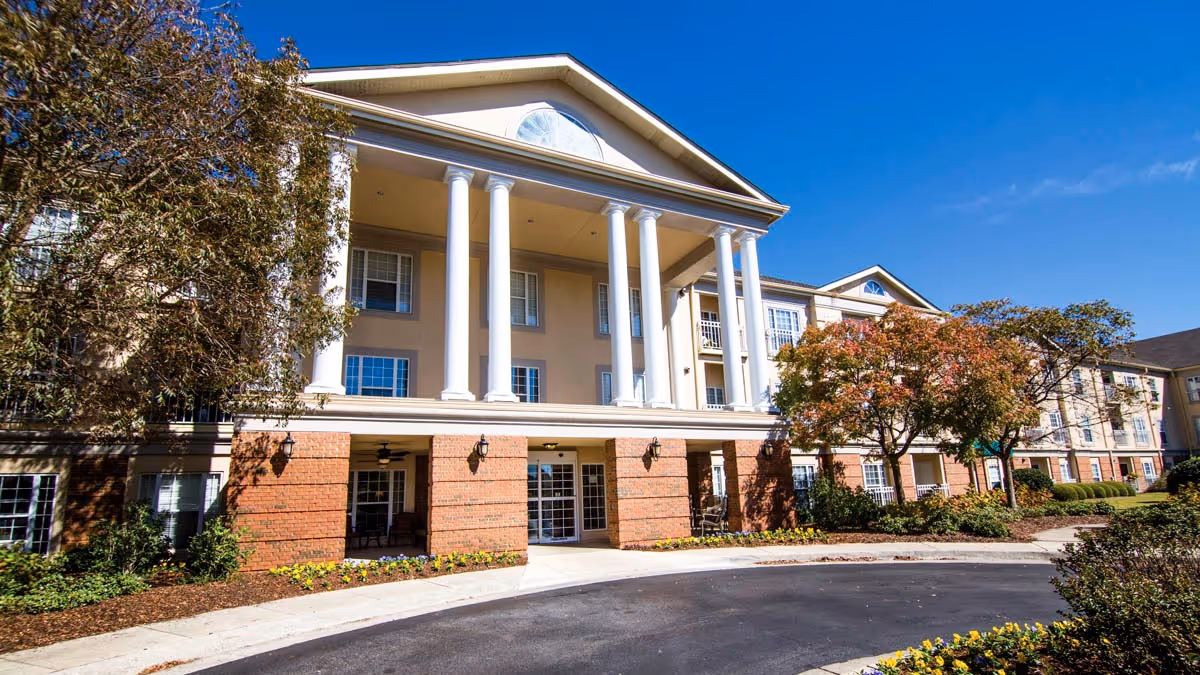Exterior view of a senior living facility building with large white columns at the entrance, brick and beige walls, surrounded by trees and landscaping under a clear blue sky.