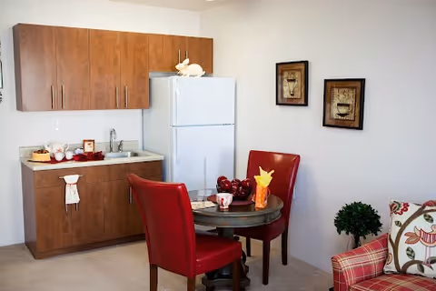A small kitchen area with wooden cabinets, a white refrigerator, and a countertop with a sink. In front of the kitchen is a round glass-top table with two red chairs. On the table are decorative items including a yellow napkin holder and a bowl of red apples. To the right, part of a patterned armchair and a small green potted plant are visible. Two framed pictures hang on the white wall above the table.