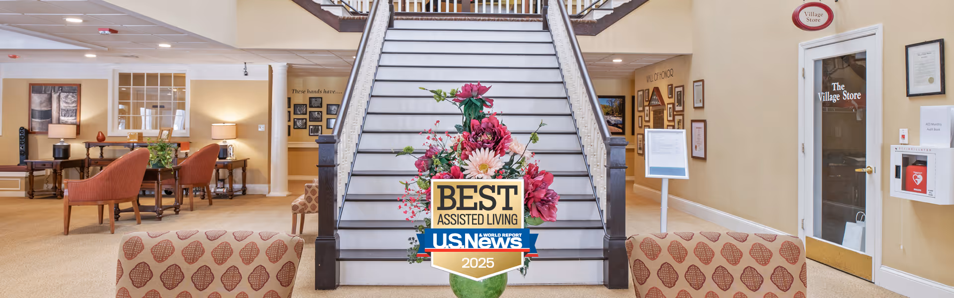 Interior view of a senior living facility lobby with a wide staircase in the center adorned with a floral arrangement. To the left, there are seating areas with chairs and tables, and to the right, a door labeled 'The Village Store'. The walls are decorated with framed pictures and certificates.