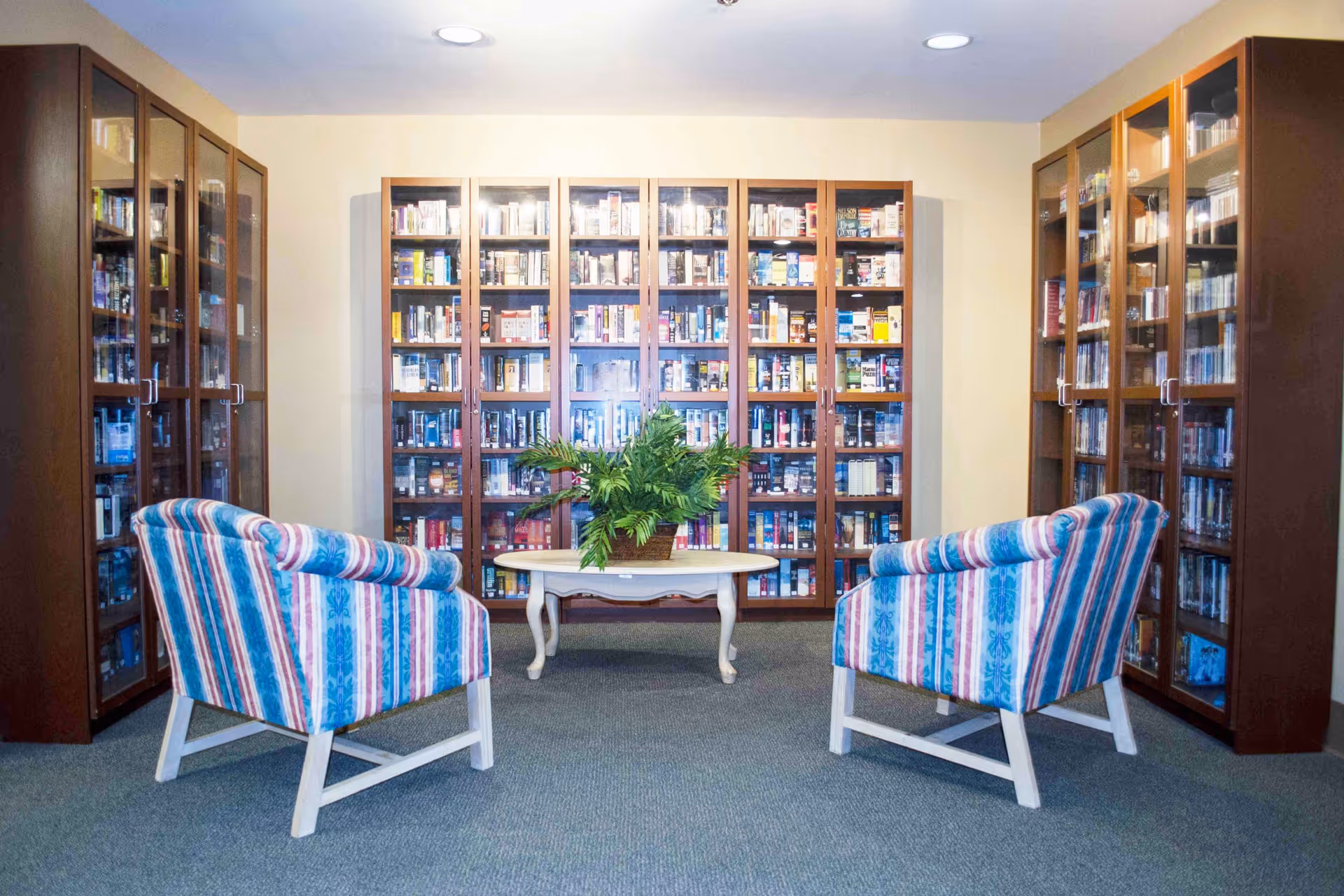 A cozy reading area in a library or common room with two striped armchairs facing a white oval table that holds a green potted plant. Behind the table are tall wooden bookshelves filled with books enclosed in glass doors.
