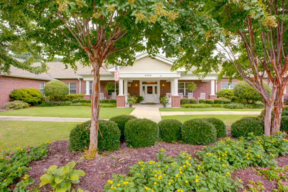 Front exterior view of a single-story brick assisted living facility with a covered entrance supported by white columns, surrounded by well-maintained landscaping including trimmed bushes, trees, and flowering plants.