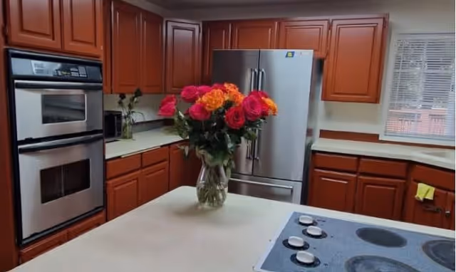 A kitchen with wooden cabinets, a stainless steel double oven, a stainless steel refrigerator, a stovetop with four burners, and a countertop island with a vase of colorful roses. There is a window with blinds on the right side.