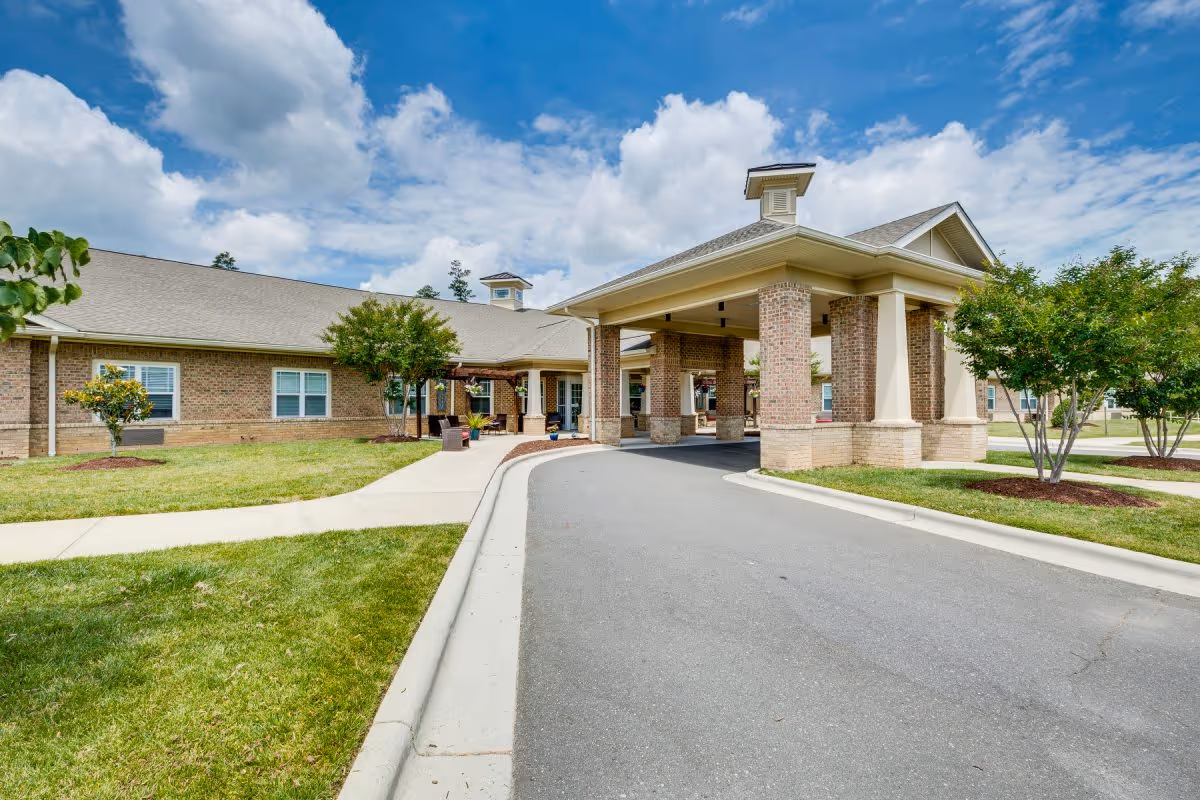 Exterior view of Chatham Ridge Assisted Living facility showing a covered entrance with brick pillars, a driveway, green lawns, small trees, and a partly cloudy blue sky.