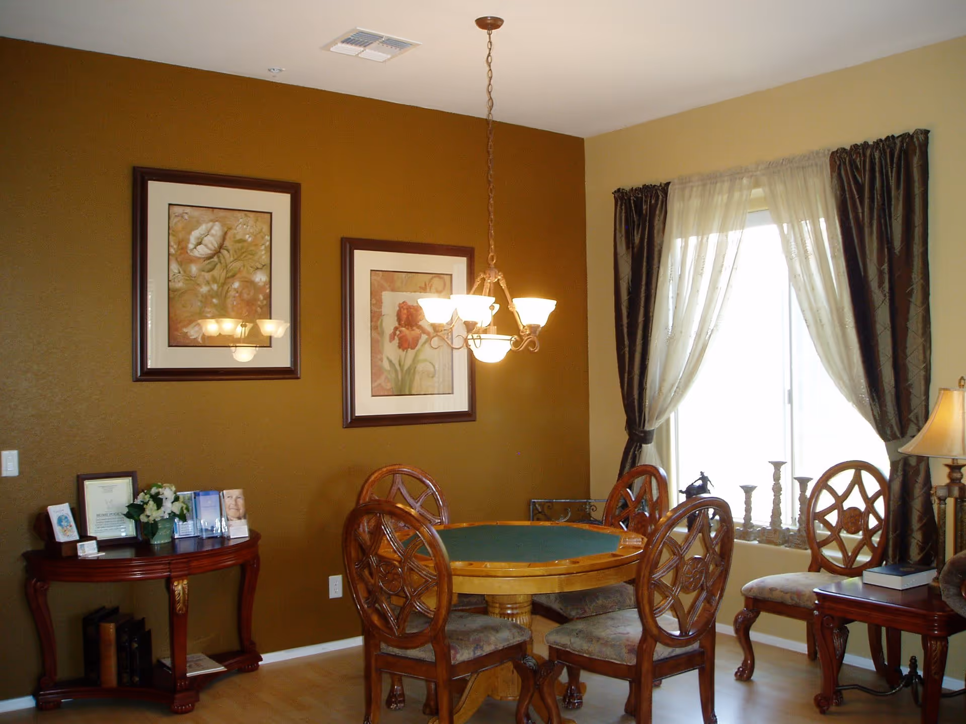 A cozy dining room with a round wooden table surrounded by four chairs with decorative backs. The walls are painted in two tones, mustard yellow and beige. Two framed floral paintings hang on the mustard yellow wall. A chandelier with six lights hangs above the table. A window with sheer white curtains and dark drapes lets in natural light. A small wooden side table with a lamp and a book is next to one of the chairs, and another small table with framed photos and brochures is against the wall.
