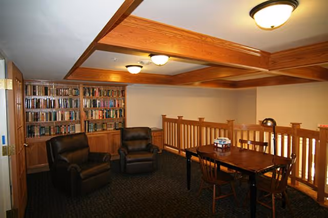 Loft-style senior living lounge with bookshelves, leather chairs, and a wooden dining table.