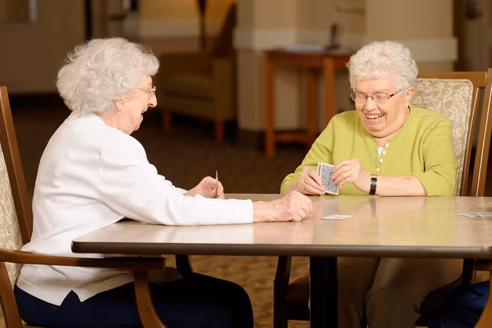 Two elderly women sitting at a table playing cards and smiling inside a well-lit room with carpeted floor and wooden furniture.