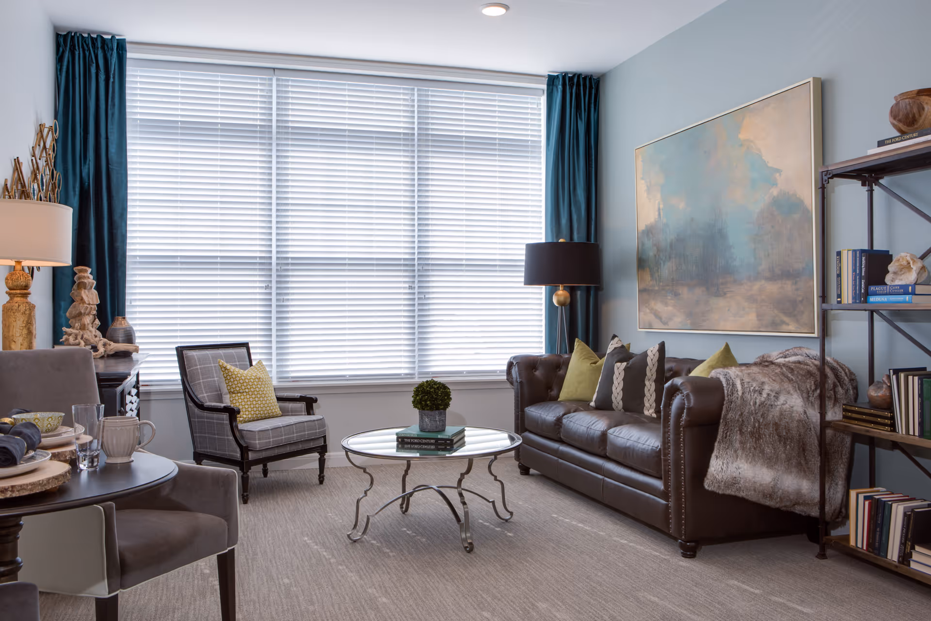 A cozy living room with a large window covered by white blinds and dark teal curtains. The room features a dark brown leather sofa with multiple decorative pillows and a fur throw, a round glass coffee table with books and a small plant, a patterned armchair with a yellow pillow, a side table with a lamp and decorative items, and a bookshelf filled with books and decor. A large abstract painting hangs on the light blue wall.